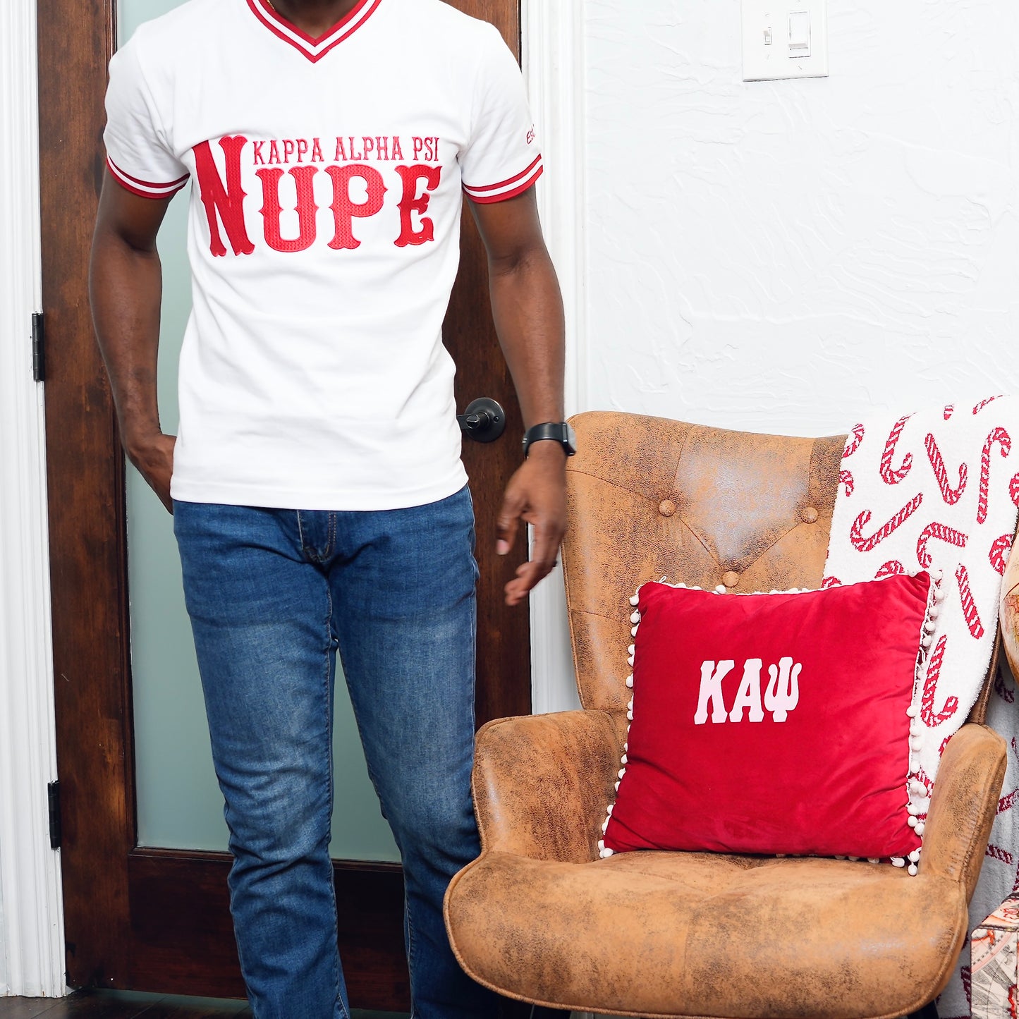 Person wearing a white t-shirt with red text standing next to a brown armchair with a red pillow featuring 'KAΨ'.The Kappa Alpha Psi "Nupe" T-shirt is a collectible item that represents the historical significance and traditions of the fraternity . It serves as a symbol of pride and belonging for members and supporters of Kappa Alpha Psi. This t-shirt is a unique piece of memorabilia that showcases the unique cultural heritage and shared values of those involved in the organization.