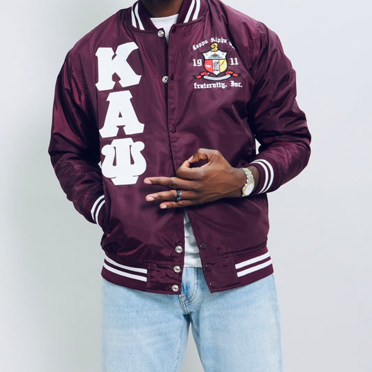 Person wearing a maroon letterman jacket with white text and logos on a plain background.Check out our Kappa Alpha Psi satin jacket selection for the very best in unique design. This Kappa Alpha Psi Baseball Nylon Jacket shows the fraternity Shield embroidered on left chest and Greek letters on right to create style and unique comfort in wear. Quality beyond imagination.

